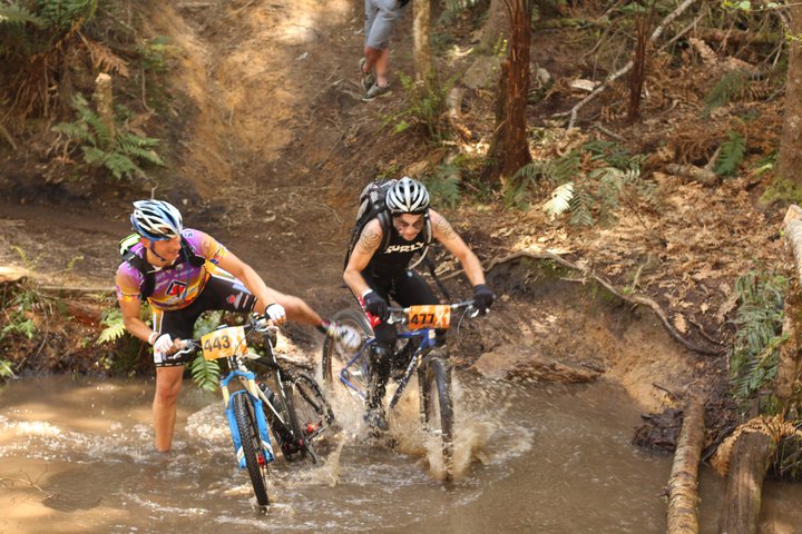 Front view of 2 cyclists riding side by side, splashing through a large mud puddle, on a trail in a forest